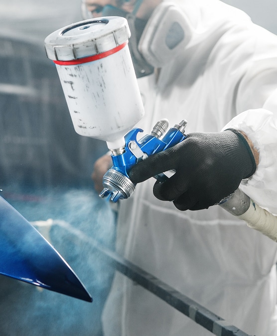 Personne en combinaison protectrice peignant une pièce de carrosserie bleue avec un pistolet à peinture dans un atelier.