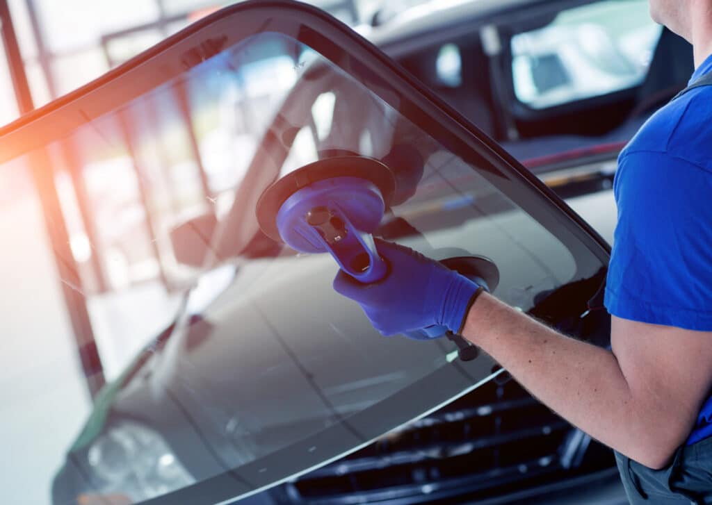 Technicien en gants bleus installe un pare-brise de voiture avec un outil à ventouses 'MADE IN GERMANY' dans un garage automobile.